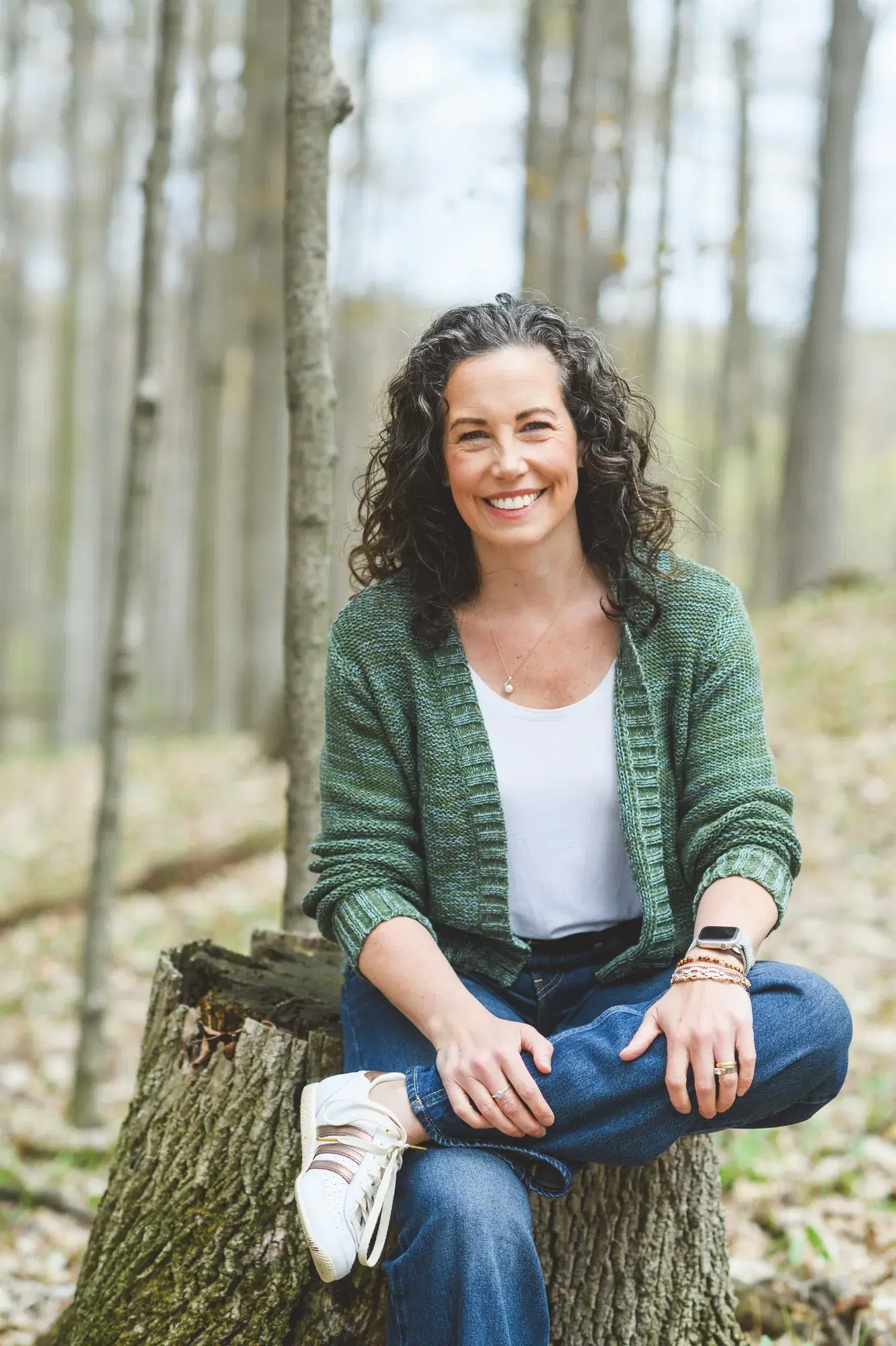 Heather sitting on a tree stump in a sunlit spring forest, wearing a sage green cardigan over a white tee, smiling toward the camera.