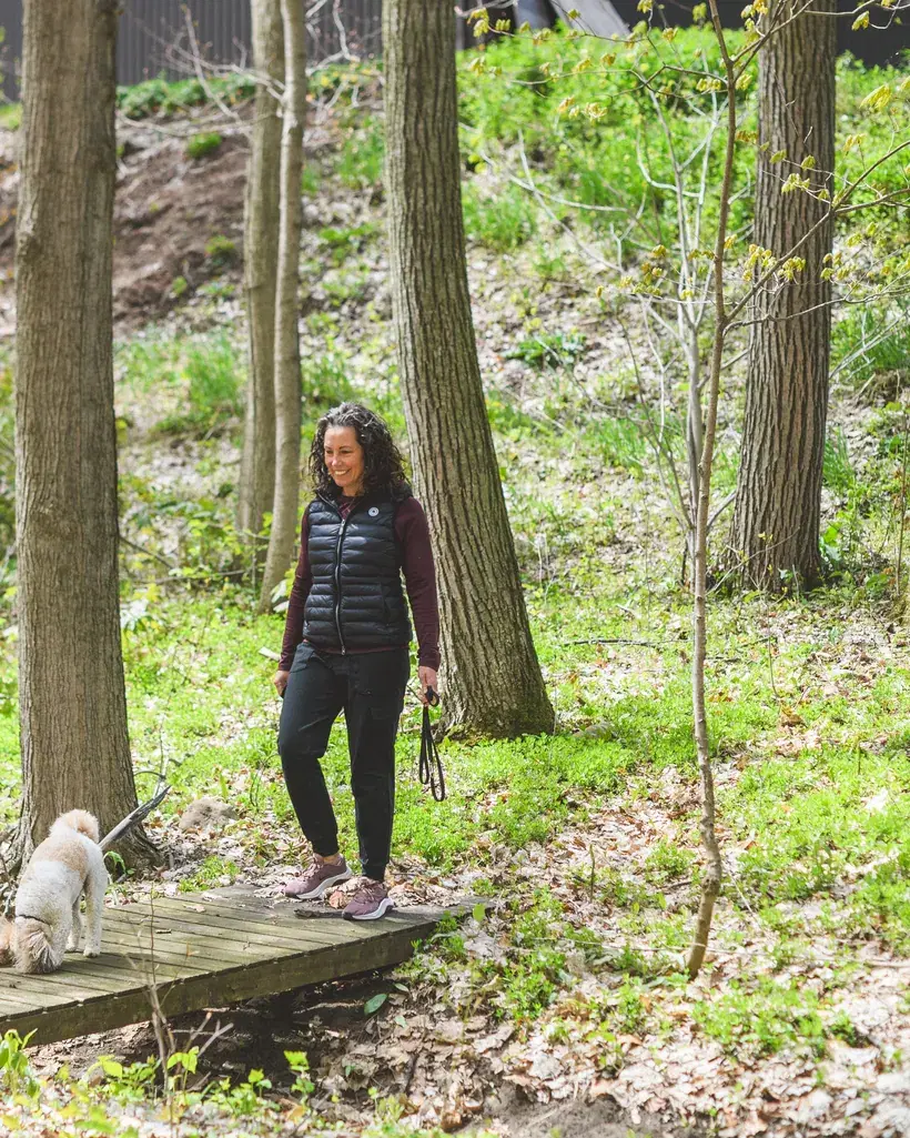 Heather walks a low wooden boardwalk through spring woodland with her small dog, smiling as she goes.