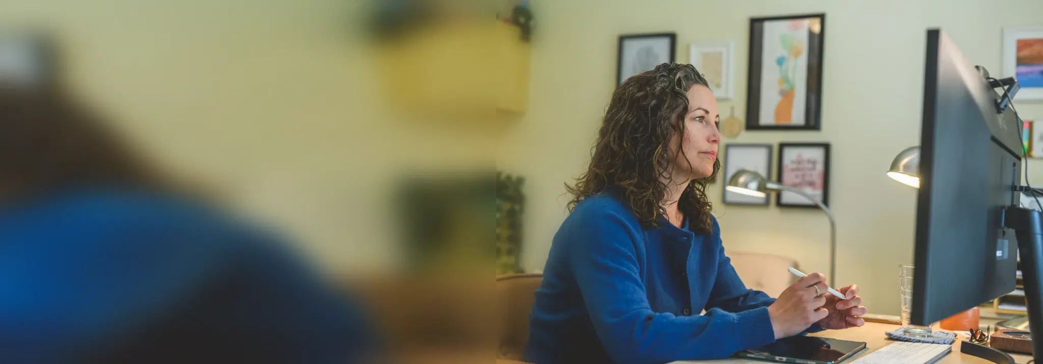 Heather Young in a blue sweater at her desk, listening attentively toward a monitor during a virtual session, framed artwork on the wall behind her.