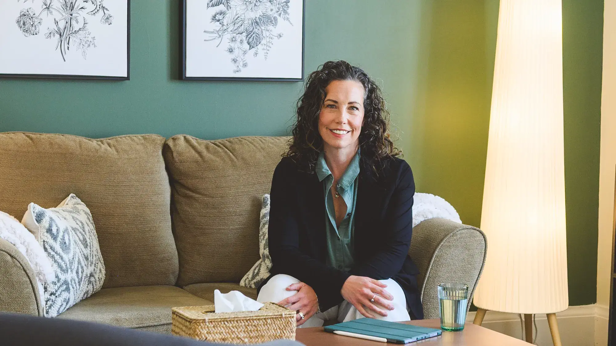 Heather Young seated on a sofa in her St. Agatha counselling office, smiling warmly, with botanical prints on a sage green wall and a soft floor lamp beside her.