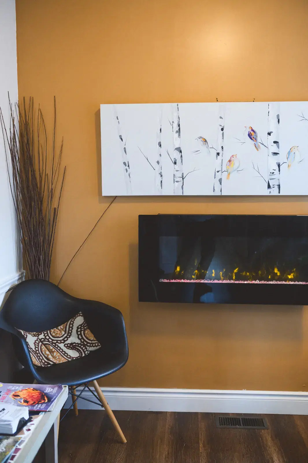 The waiting area: a black Eames chair with a patterned cushion in front of an electric fireplace and a canvas painting of birds on birch trees against an ochre wall.