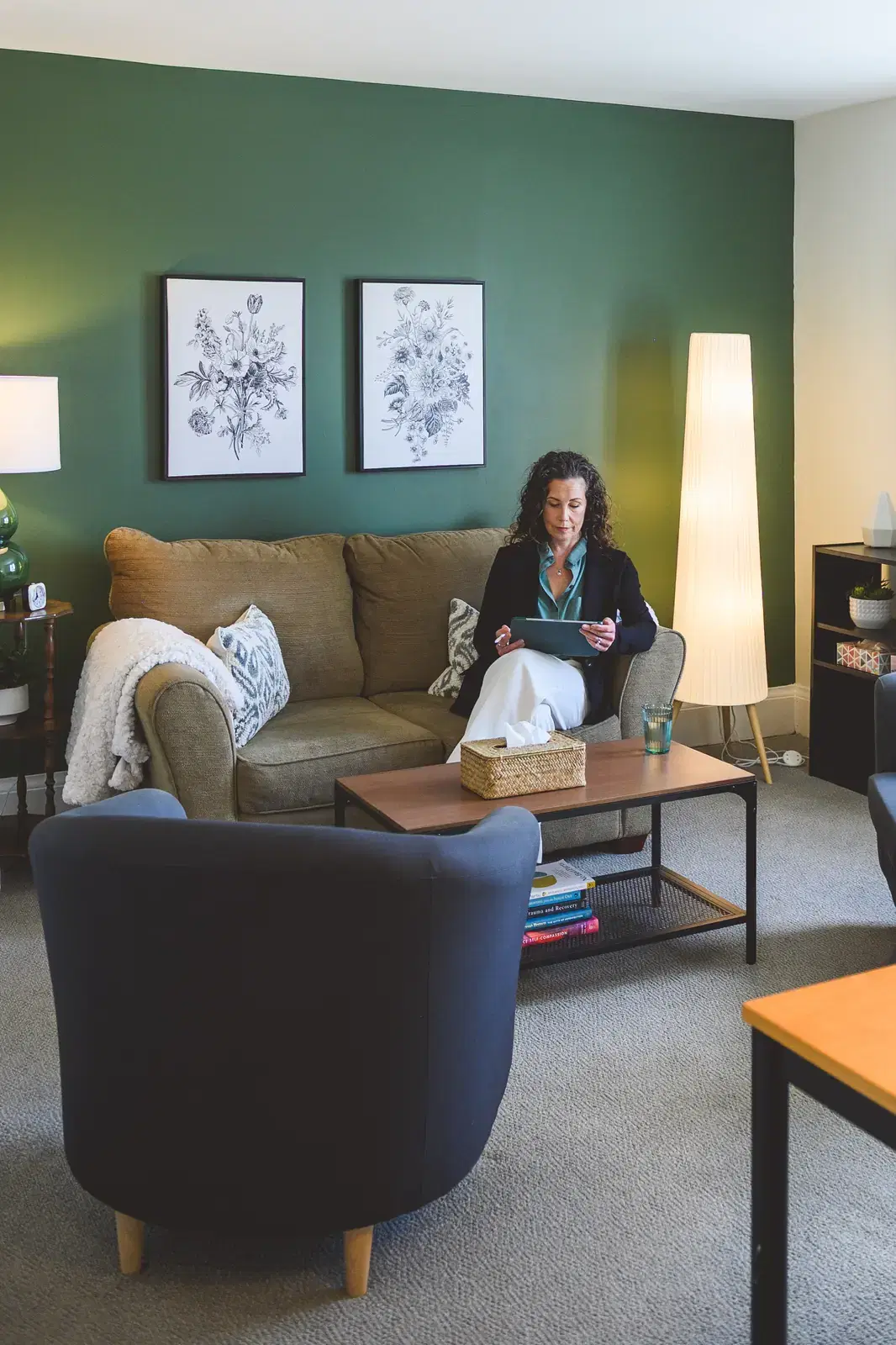 The office from the client's seat: a beige sofa and arm chair facing each other across a wooden coffee table, with botanical prints on a sage green accent wall.