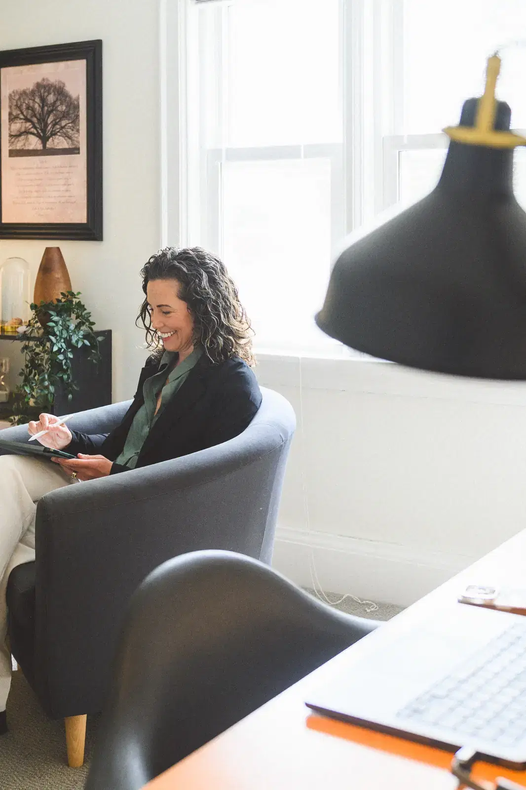 Heather seated in the office chair holding a tablet, smiling, with a desk lamp in the foreground evoking an in-session moment.