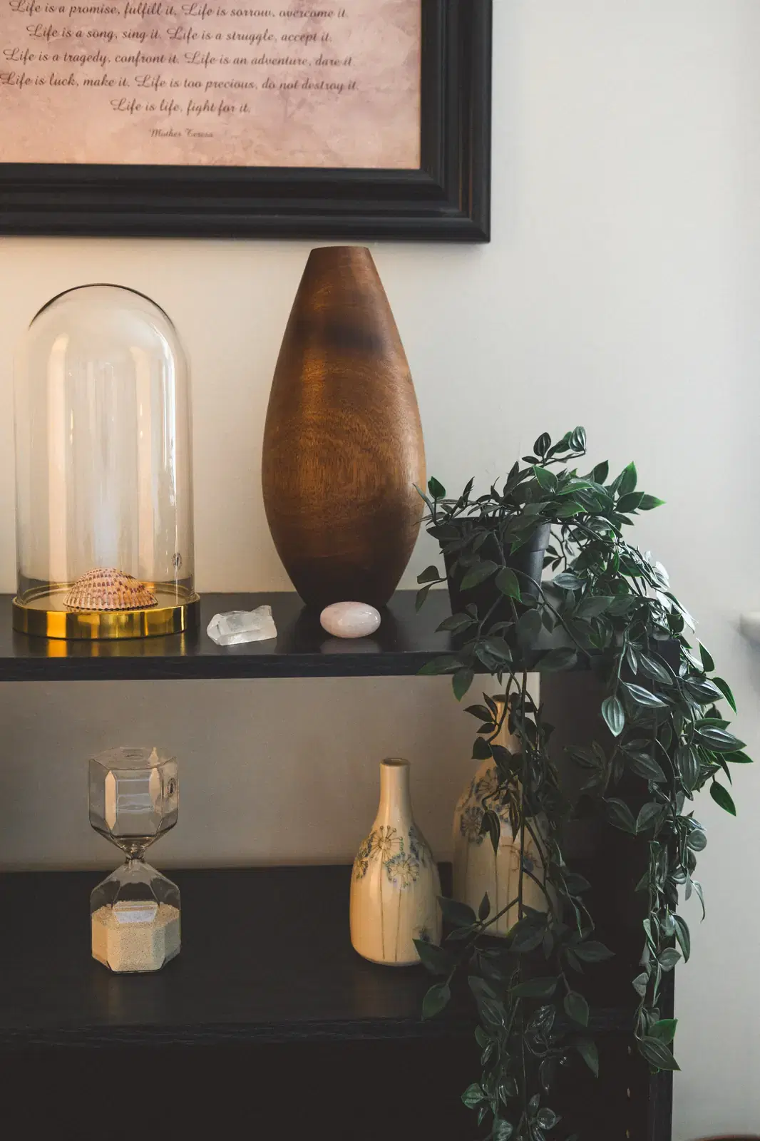 A black bookshelf beside the office sofa, holding a wooden vase, glass dome with a seashell, crystals, an hourglass, and trailing greenery, beneath a framed Mother Teresa quote.