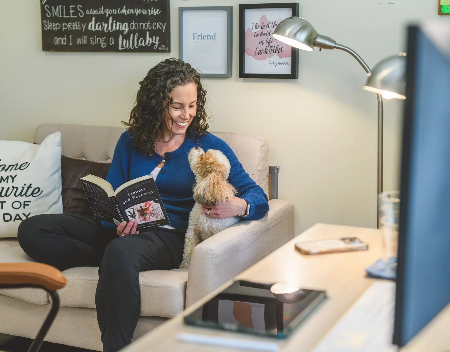 Heather Young sits on a sofa in a blue sweater, smiling down at her small curly-haired dog as she reads, framed prints on the wall behind her.
