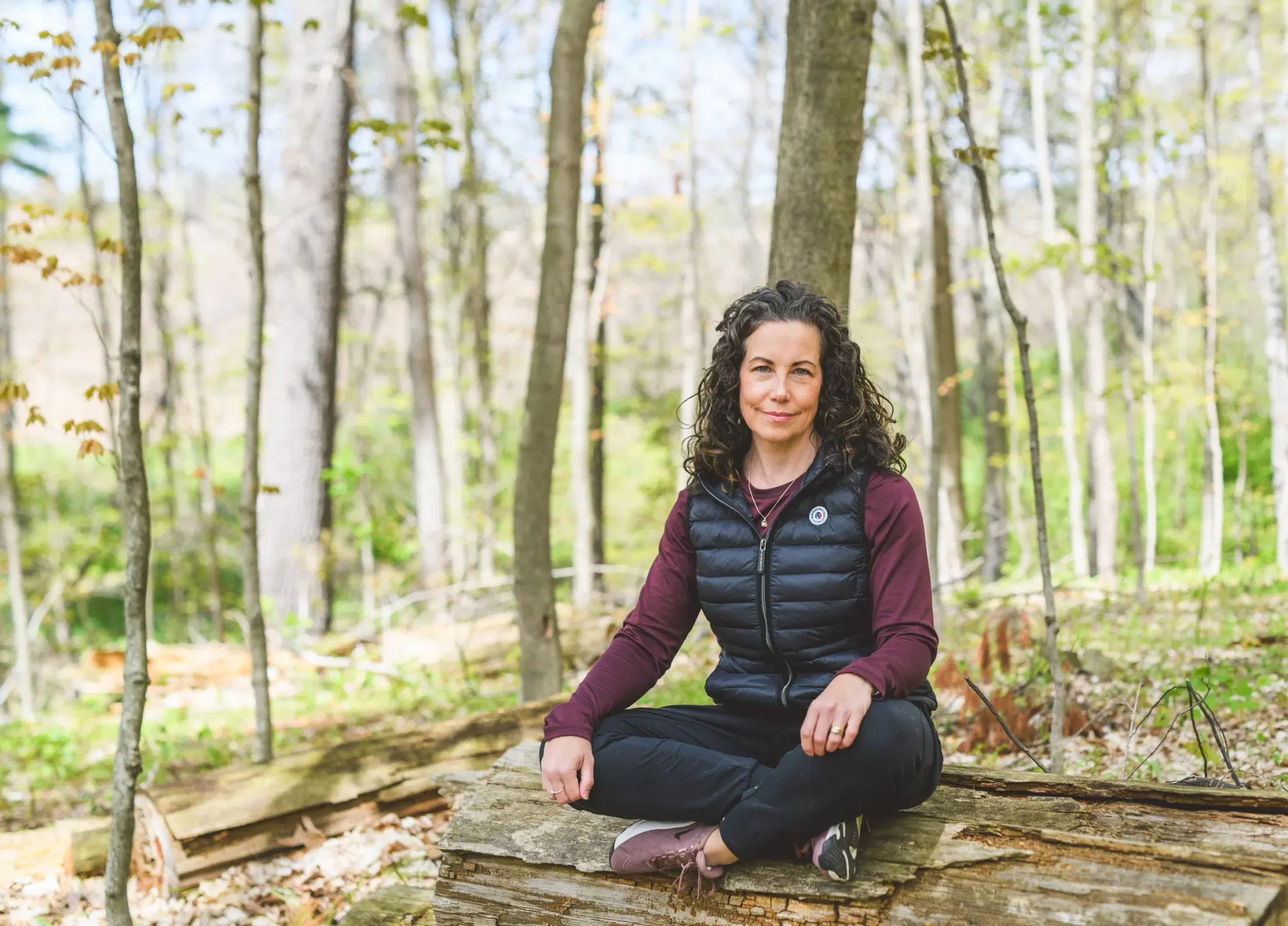 Heather Young sits cross-legged on a fallen log in a spring forest, looking calmly at the camera, surrounded by young trees and bright new leaves.