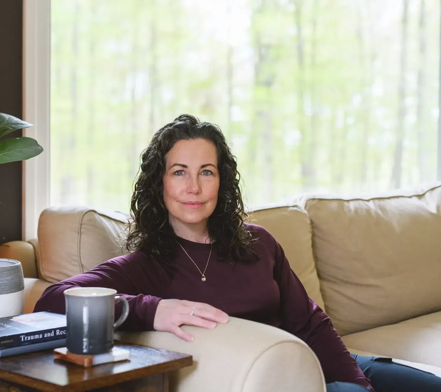 Heather sits on a cream sofa beside a side table with a plant, a mug, and a book, looking calmly toward the camera with forest visible through the window behind her.