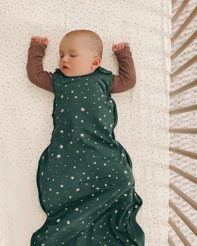 A baby sleeps on their back in a bare crib, wearing a sleep sack, on a firm mattress with a fitted sheet and nothing else in the crib.
