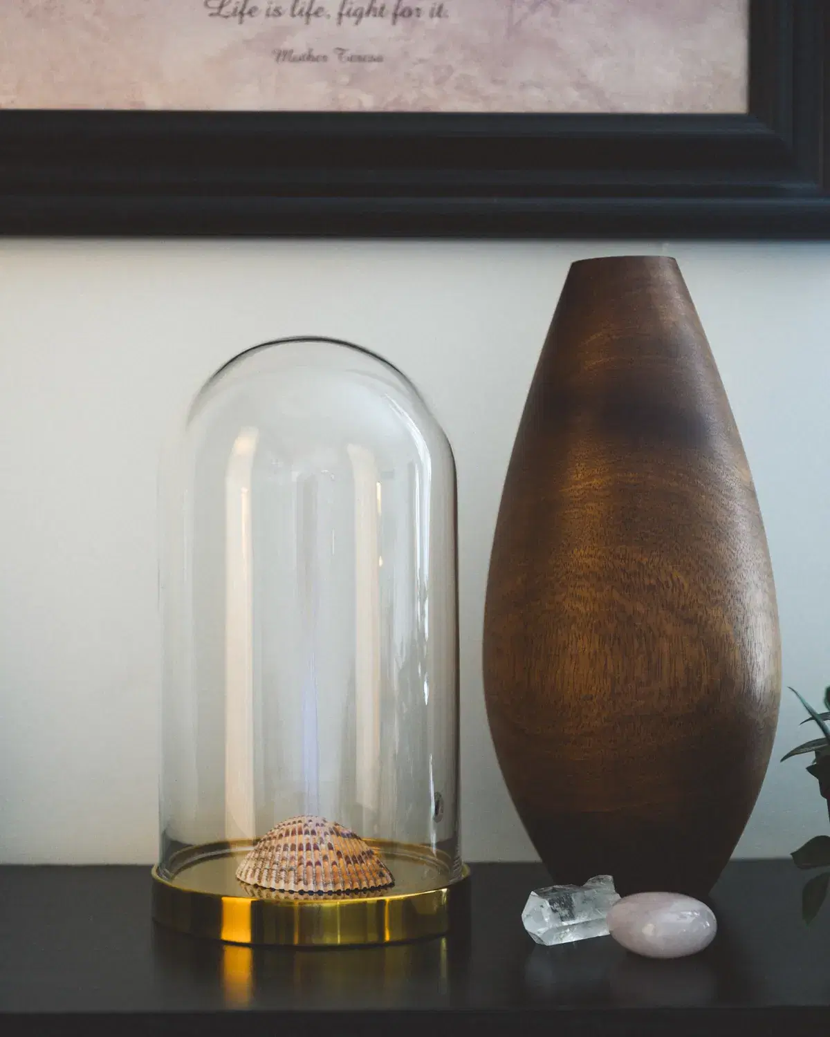 A glass cloche and a smooth wooden vessel resting on a dark shelf in still, quiet daylight.