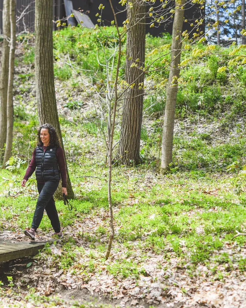 Heather walks toward the camera along a low boardwalk through dappled spring woodland.
