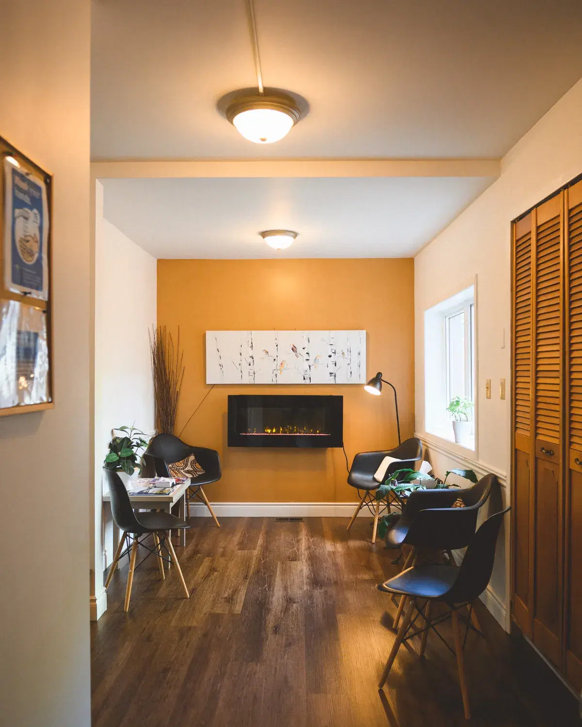 A calm interior looking through toward a lit fireplace and seating area, warm daylight falling across a wooden floor.
