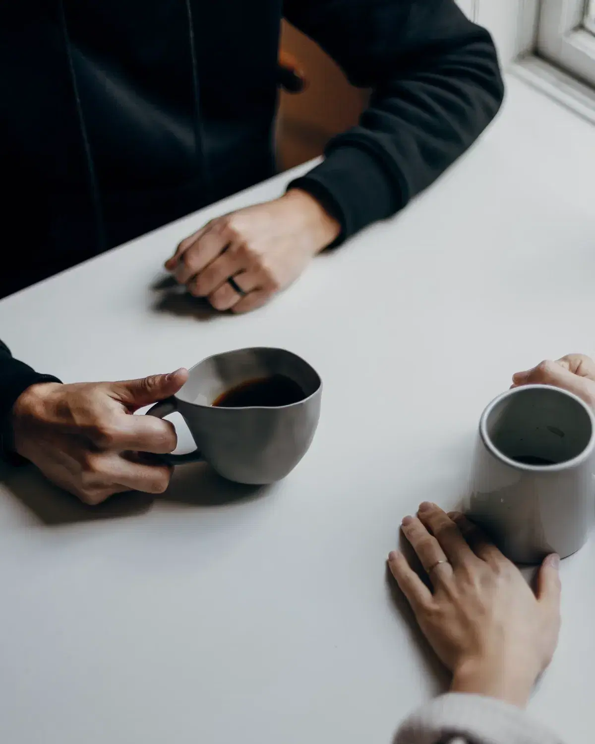Two people sit across a table from one another with mugs of coffee, hands resting calmly, in soft natural light.