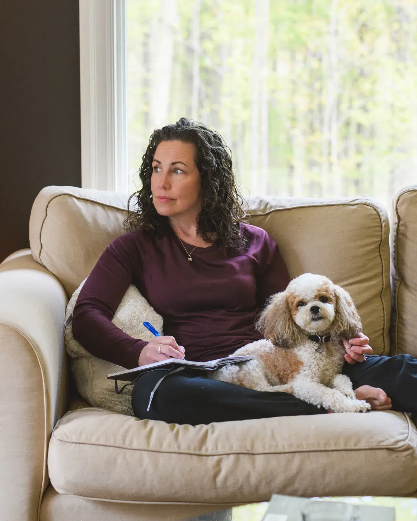 Heather sits in a cream armchair beside a bright window with an open notebook and her small dog resting beside her, spring woodland visible outside.
